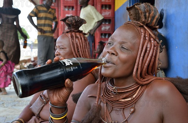 Traditionally dressed Himba woman drinking a coke in front of a bar, Opuwo, Kaokoland, Kunene, Namibia, Africa