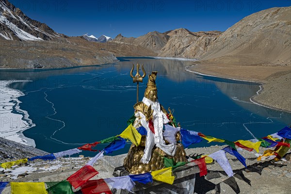 Tilicho Lake covered with ice in front of mountain scenery with Buddhist prayer flags and Hindu Shiva statue