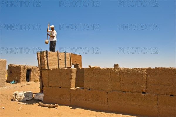 Elderly man wearing a white turban using a wooden tool to compact clay into a wooden casing for the construction of a wall made of rammed earth
