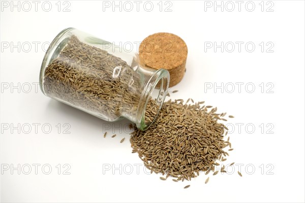 Cumin seeds spilling out of a 150ml glass spice jar against a white background