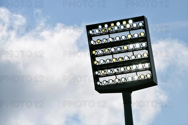 Floodlight mast switched on in front of blue sky with clouds