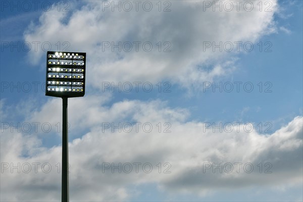 Floodlight mast switched on in front of blue sky with clouds