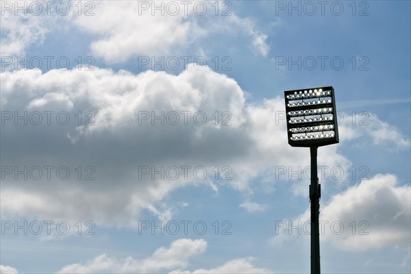 Floodlight mast switched on in front of blue sky with clouds