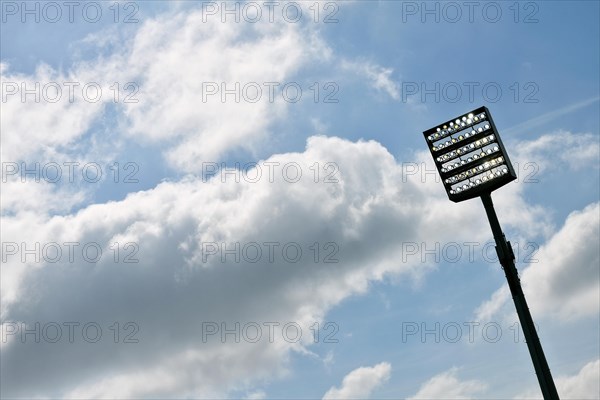 Floodlight mast switched on in front of blue sky with clouds