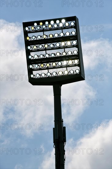 Floodlight mast switched on in front of blue sky with clouds