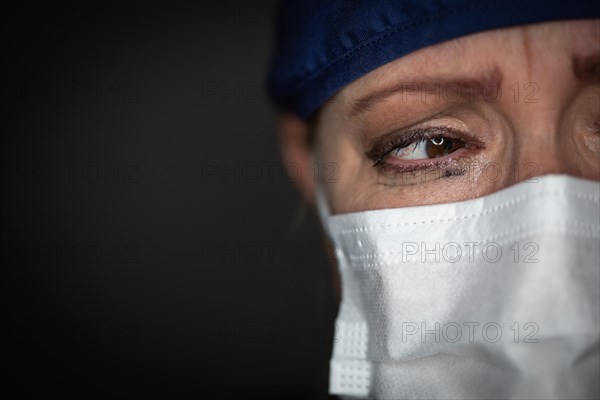 Tearful stressed female doctor or nurse wearing medical face mask on dark background