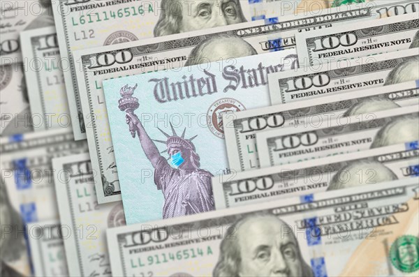 United states IRS stimulus check with statue of liberty wearing medical face mask resting on hundred dollar bills