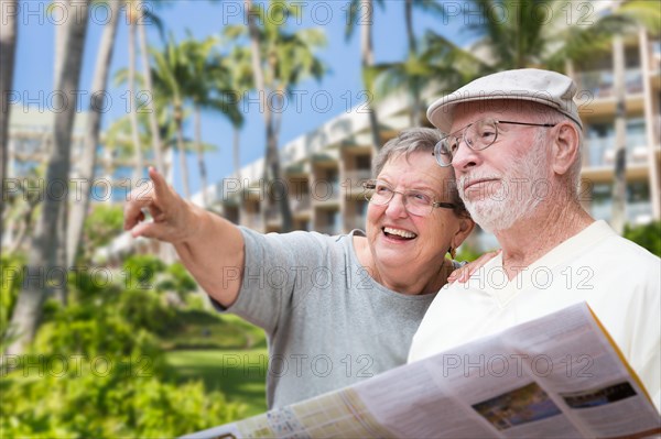 Happy senior adult couple tourists with brochure next to tropical hotel and palm trees