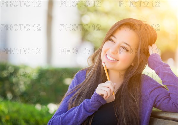 Attractive pensive mixed race female student with pencil sitting on campus bench