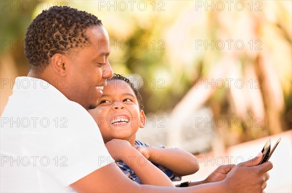 African american father and mixed race son using computer tablet on bench in park