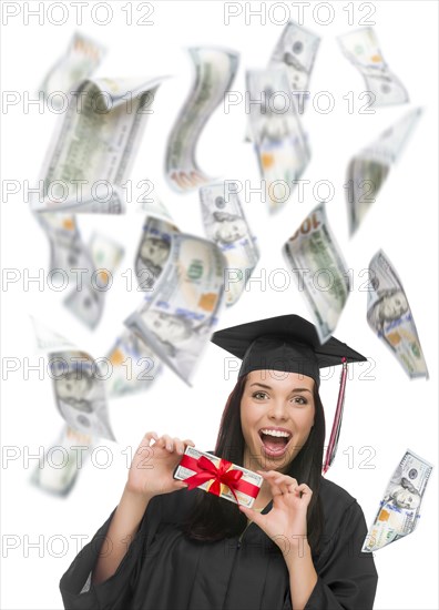 Excited female graduate in cap and gown holding stack of $100 bills with many falling around her on white