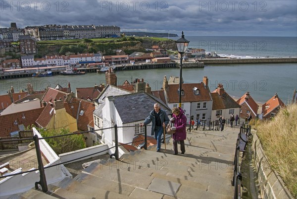 Holiday makers walking up steps in seaside town to Whitby Abbey on clifftop