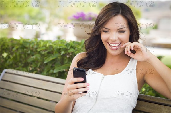 Attractive smiling young adult female texting on cell phone outdoors on a bench