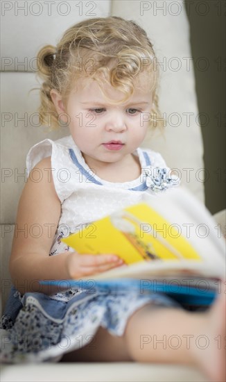 Adorable blonde haired blue eyed little girl reading her book in the chair