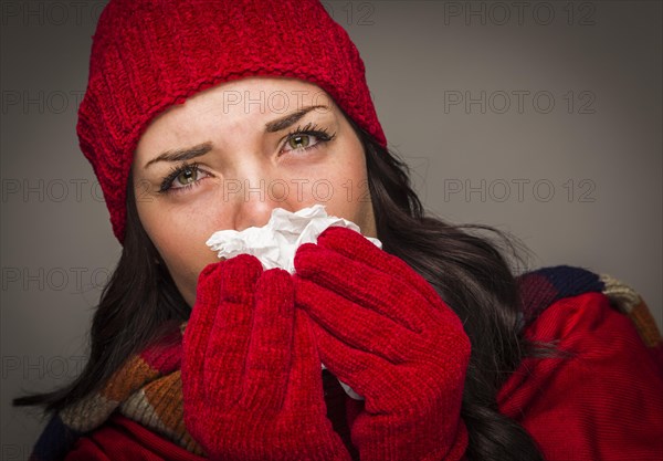 Sick mixed-race woman wearing winter hat and gloves blowing her sore nose with a tissue