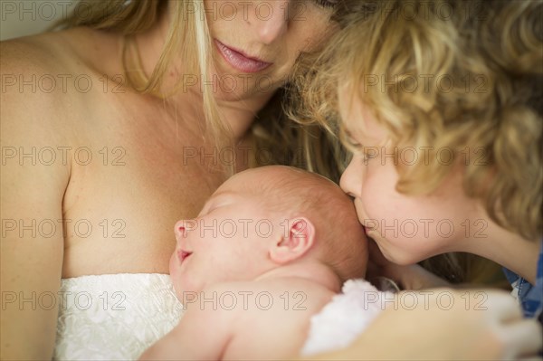 Beautiful young mother holds newborn baby girl as brother kisses her head