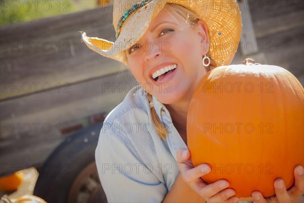 Beautiful blond female rancher wearing cowboy hat holds a pumpkin in a rustic country setting