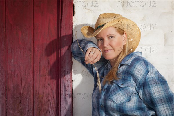 Beautiful cowgirl wearing cowboy hat leaning against old adobe wall and red door