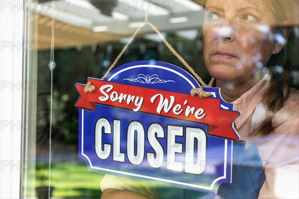 Sad female store owner turning sign to closed in window
