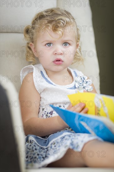 Adorable blonde haired blue eyed little girl reading her book in the chair