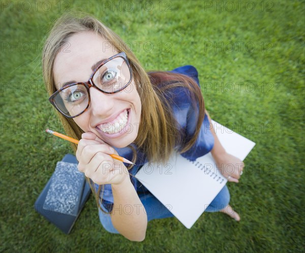 Fun wide angle portrait of pretty young woman with books and pencil sitting in the grass outdoors