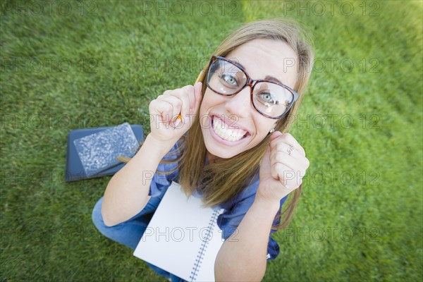 Fun wide angle portrait of pretty young woman with books and pencil sitting in the grass outdoors