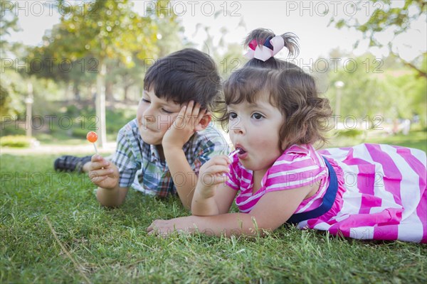 Cute young brother and baby sister enjoying their lollipops outdoors on the grass