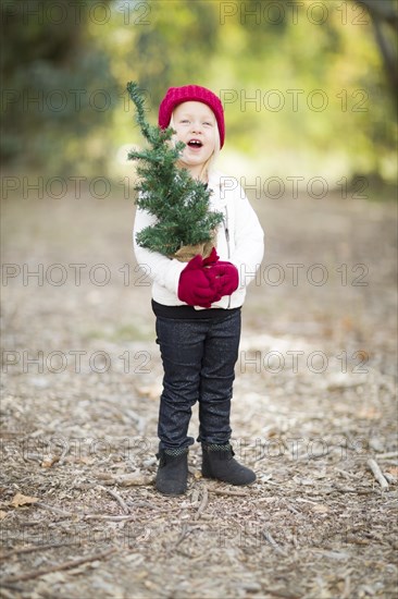 Baby girl in red mittens and cap holding small christmas tree outdoors