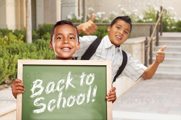 Happy hispanic boys giving thumbs up holding back to school chalk board outside on school campus