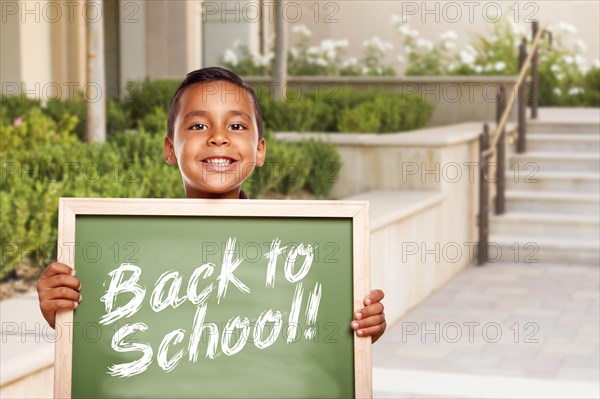 Happy hispanic boy holding back to school chalk board outside on school campus