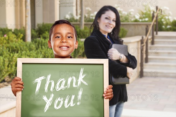 Happy hispanic boy holding thank you chalk board outside on school campus as teacher looks on