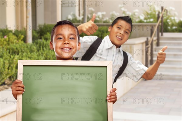 Happy hispanic boys with thumbs up holding blank chalk board outside on school campus