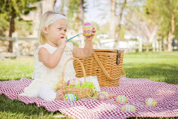 Cute baby girl enjoys coloring her easter eggs on picnic blanket in the grass