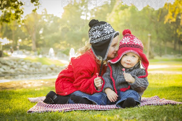 Little girl whispers A secret to her baby brother wearing winter coats and hats sitting outdoors at the park