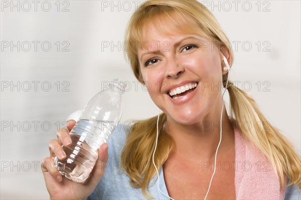 Pretty blonde woman with towel and ear phones drinking from water bottle in her kitchen