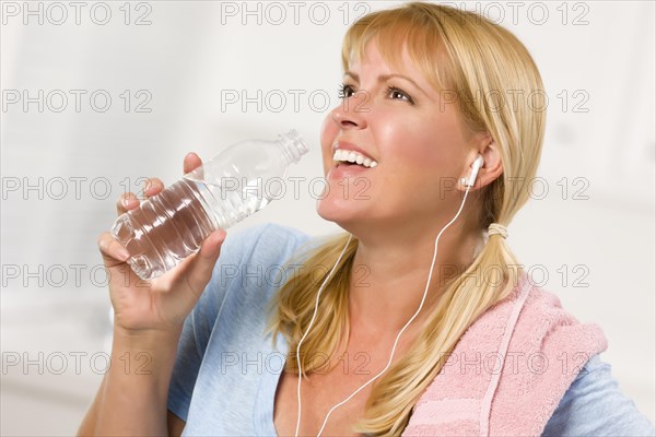 Pretty blonde woman with towel and ear phones drinking from water bottle in her kitchen