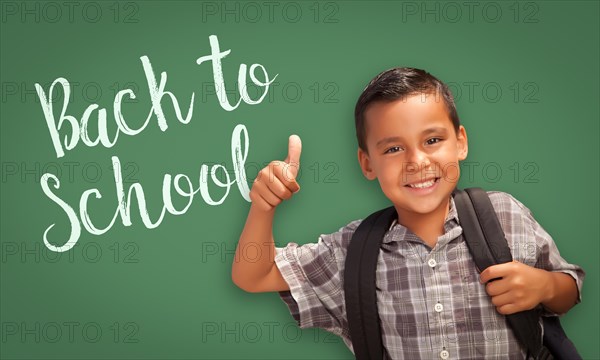 Cute hispanic boy with thumbs up wearing A backpack in front of chalk board with back to school written on it