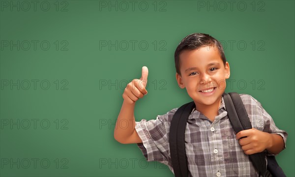 Cute hispanic boy with thumbs up wearing backpack in front of blank chalk board