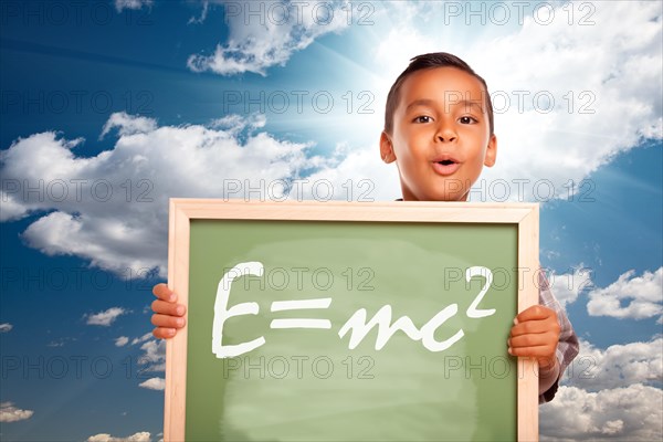 Proud hispanic boy holding chalkboard with theory of relativity over blue sky and sun burst clouds