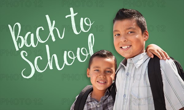 Cute hispanic boys wearing backpacks in front of back to school written on chalk board