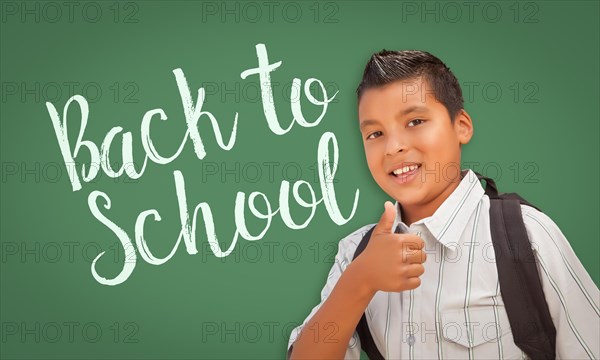 Cute hispanic boy with thumbs up wearing A backpack in front of chalk board with back to school written on it