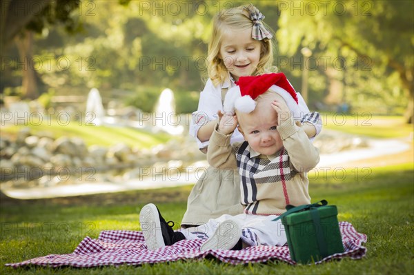 Sweet little girl tries to put A santa hat on her reluctant baby brother outdoors at the park