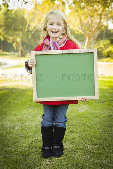 Cute little blonde wearing winter coat and scarf holding a green chalkboard outdoors