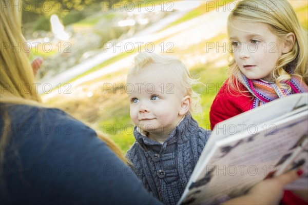 Mother reading a book to her two adorable blonde children wearing winter coats outdoors