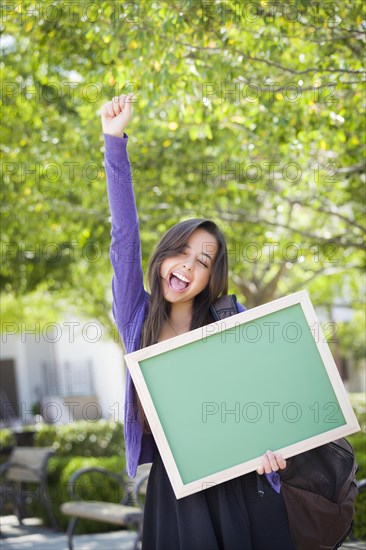 Portrait of an attractive excited mixed-race female student holding blank chalkboard and carrying backpack on school campus