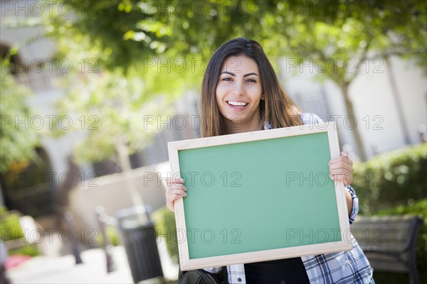 Portrait of an attractive excited mixed-race female student holding blank chalkboard and carrying backpack on school campus