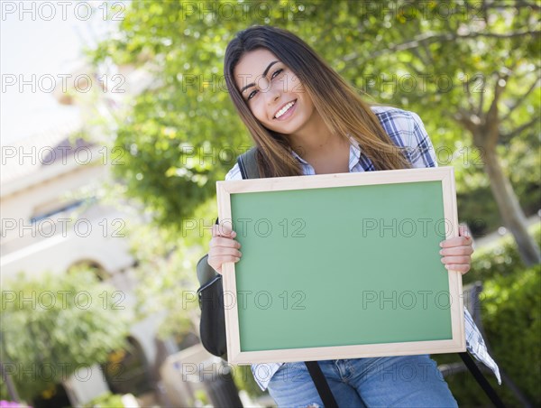 Portrait of an attractive excited mixed-race female student holding blank chalkboard and carrying backpack on school campus