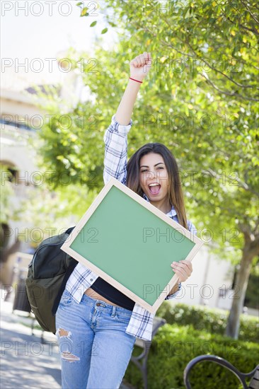 Portrait of an attractive excited mixed-race female student holding blank chalkboard and carrying backpack on school campus