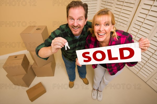 Goofy couple holding key and sold sign in room with packed cardboard boxes