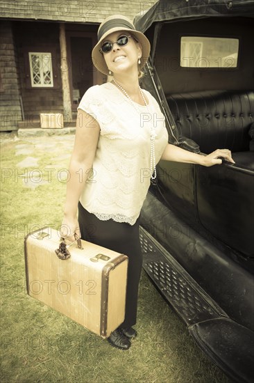 Happy 1920s dressed girl holding suitcase next to vintage car and cabin